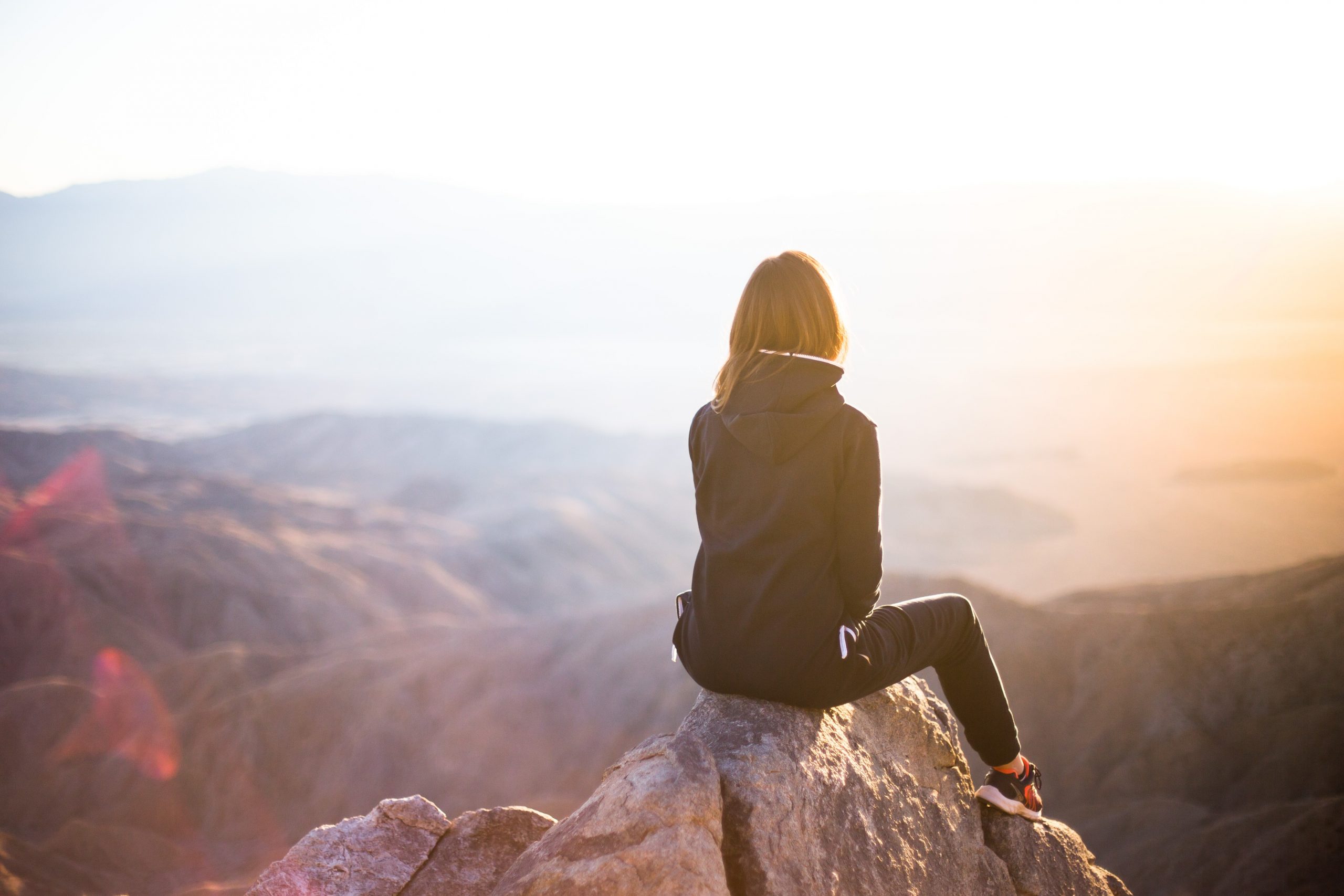 Girl Sitting with her Back Turned on a Rock on top of a Mountain Looking At The Valley