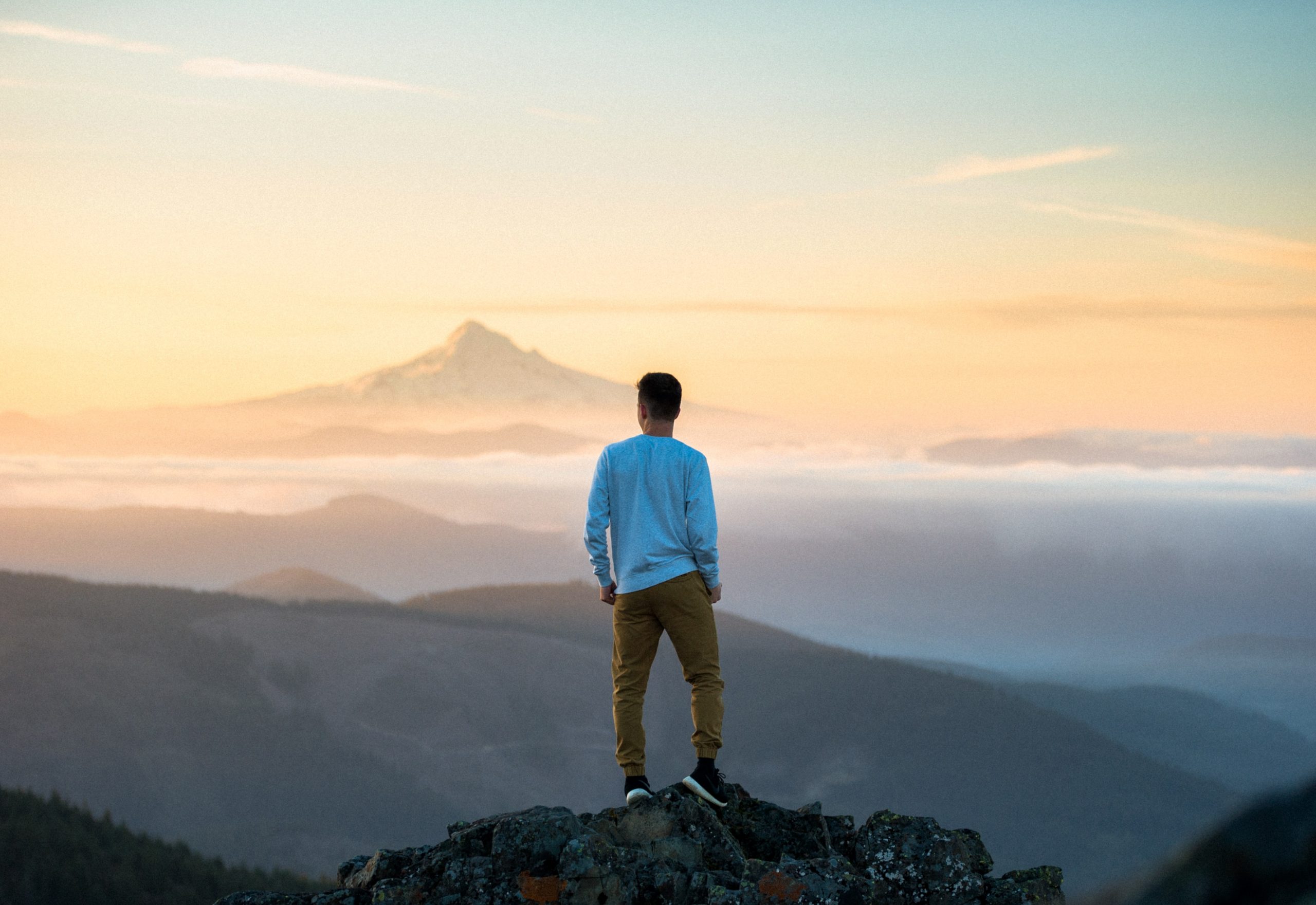 Man Standing with his Back Turned on a Rock on top of a Mountain Looking At The Valley